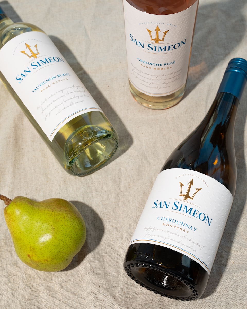 Three bottles of San Simeon wine and a green pear arranged on a light-colored fabric surface.