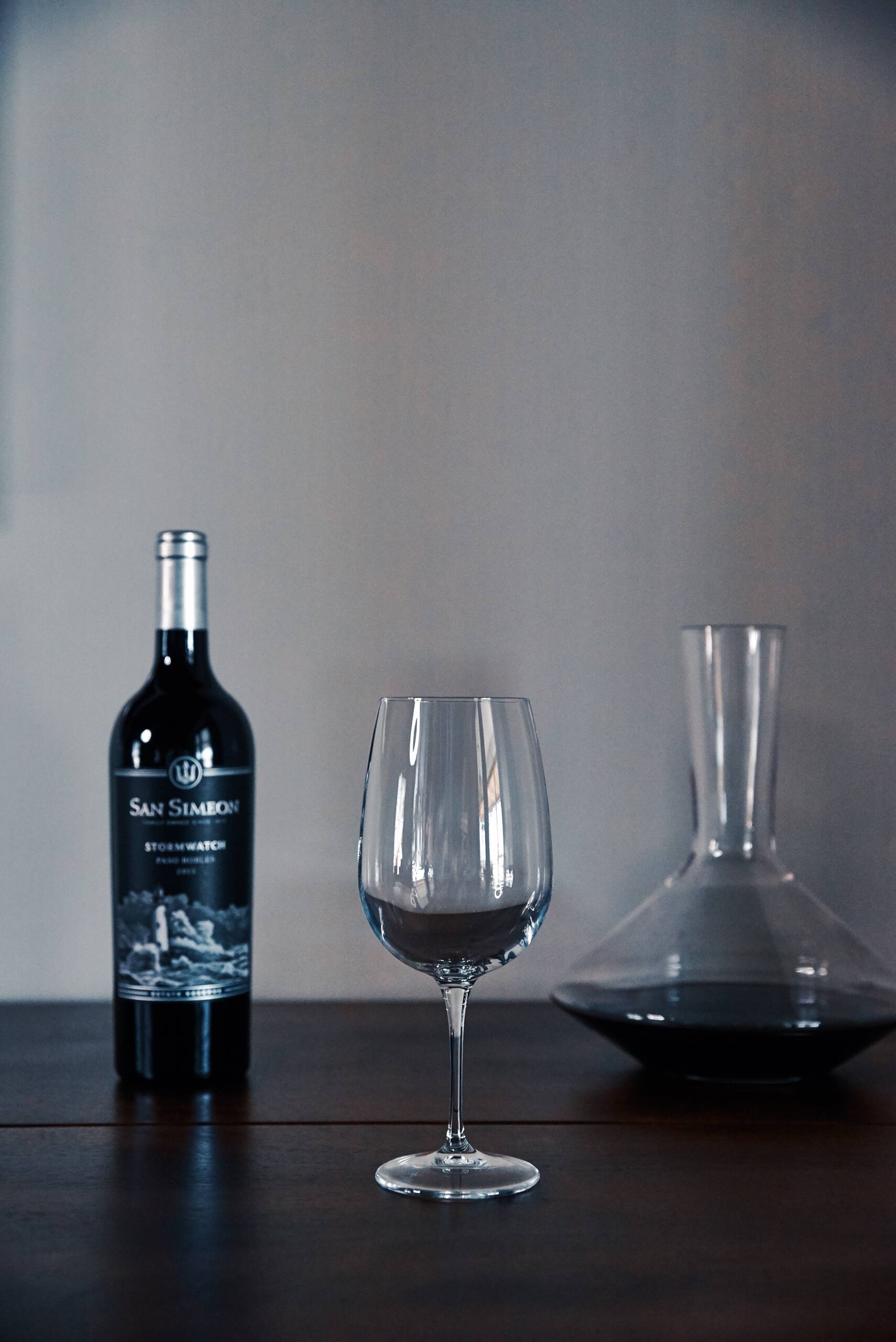 A bottle of San Simeon wine, an empty wine glass, and a glass decanter with red wine are arranged on a wooden surface against a plain background.